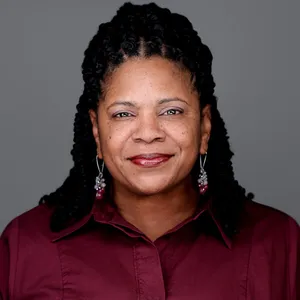Corporate headshot of African-American woman with long dark braided hairstyle wearing burgundy collared button-down shirt and silver dangle earrings photographed against gray studio background with even lighting by headshot photographer Richard Waine