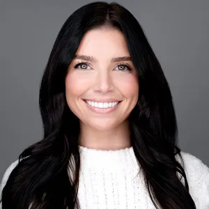 Corporate headshot of caucasian woman with long dark wavy hair and bright smile wearing white cable-knit sweater photographed against medium gray background with clean even lighting by Richard Waine