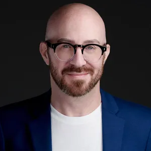 Professional linkedin profile headshot of bald man with reddish-brown beard and eyeglasses wearing navy blue blazer over white crew neck t-shirt photographed against dark charcoal studio background with moody dramatic lighting by Richard Waine