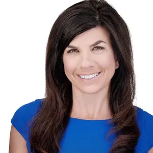 Professional executive headshot of a caucasian woman with long layered dark brown hair styled with side-swept bangs and warm smile wearing vibrant royal blue cap sleeve sheath dress photographed against seamless white studio backdrop with high-key professional lighting by Lancaster Pennsylvania headshot photographer Richard Waine