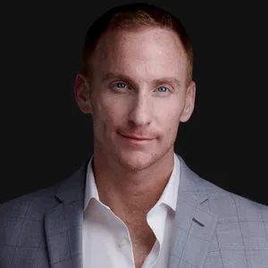 Business portrait of caucasian man with short reddish-brown hair and matching mustache wearing light gray textured suit jacket over white open-collar dress shirt photographed against dark gray studio backdrop with dramatic lighting by headshot photographer Richard Waine