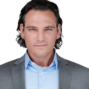 Executive portrait of caucasian man with slicked-back dark wavy hair wearing charcoal gray textured suit jacket over light blue dress shirt on white background photographed by Richard Waine