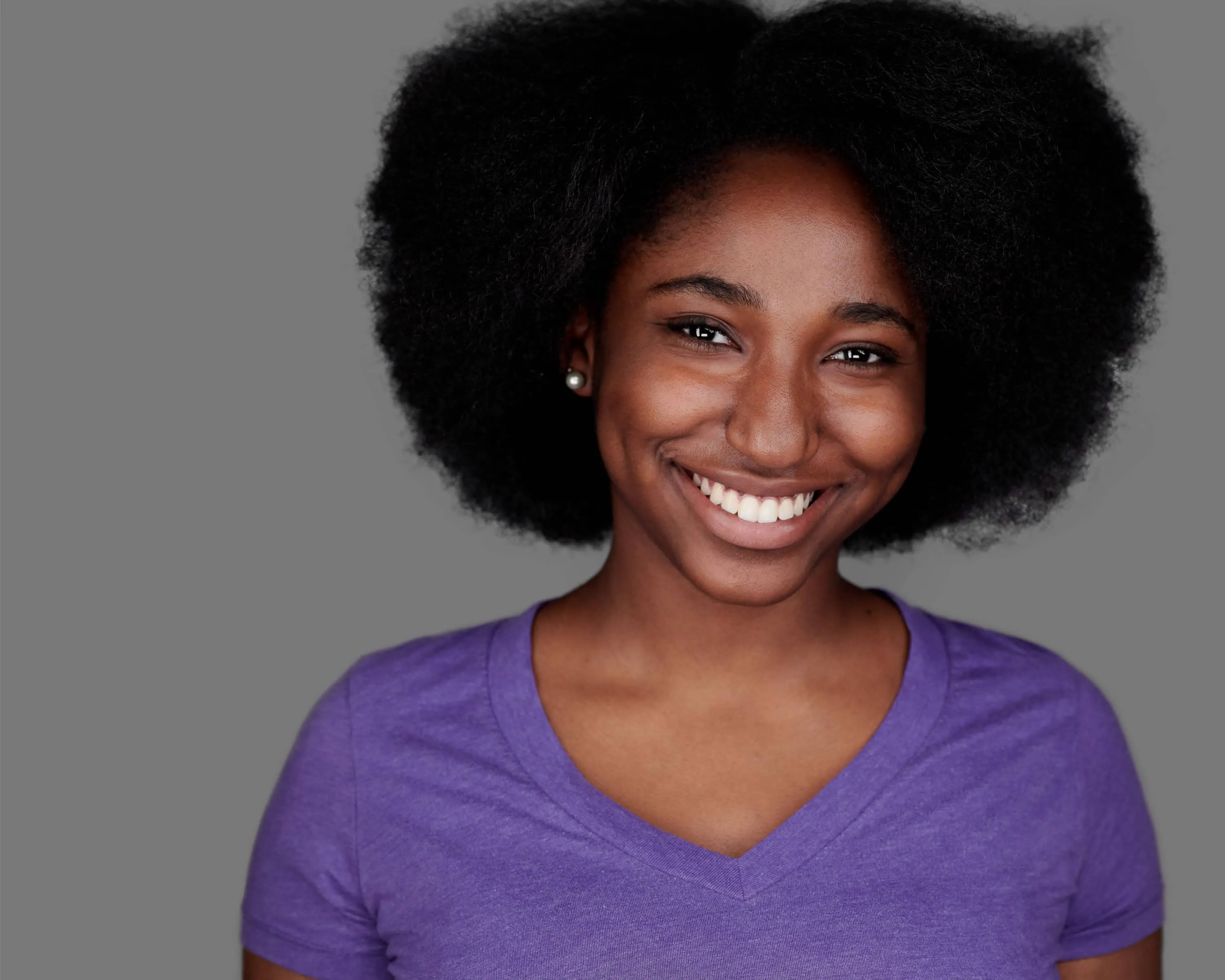 Young Black woman with voluminous natural afro wearing purple v-neck, genuine warm smile with pearl stud earrings, gray background