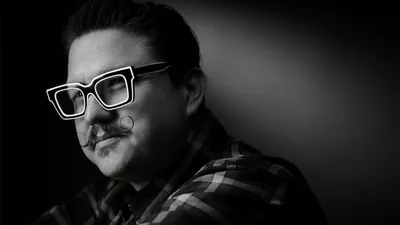 Black and white artistic portrait of Las Vegas photographer Mondo Rojas with slicked-back short dark hair wearing oversized white-framed square eyeglasses with styled handlebar mustache in checkered flannel shirt photographed with dramatic side lighting against dark gradient background