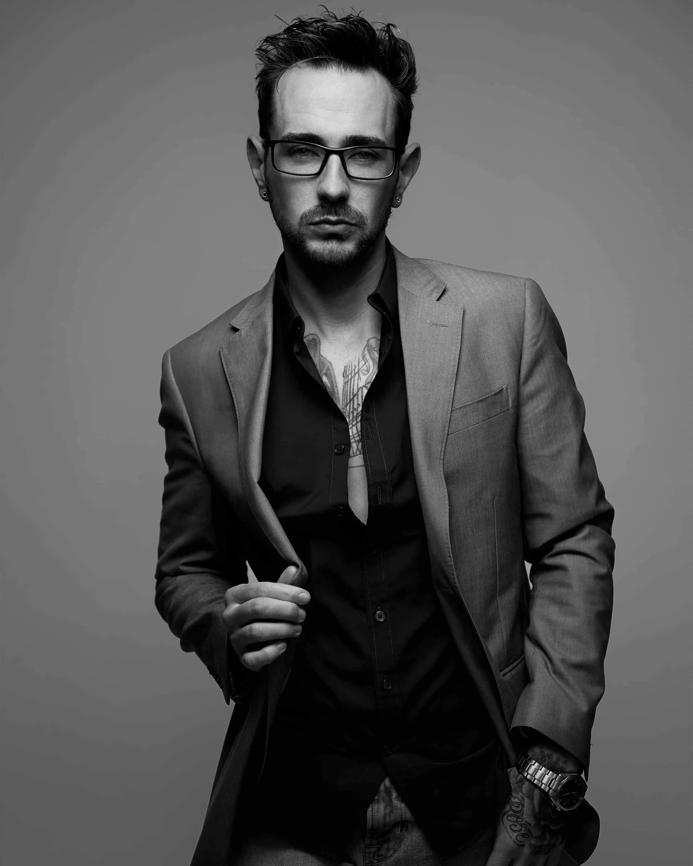 Black and white portrait of male model with styled dark hair and goatee wearing rectangular frame eyeglasses gray suit jacket over black vest and open-collar shirt with visible wrist watch, arm and wrist tattoo and chest tattoo photographed against gray studio background by Richard Waine Photography Lancaster PA, model portrait