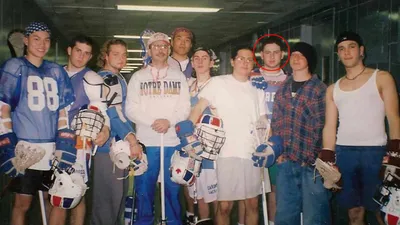 Vintage 1996 team photograph of nine Cardozo High School varsity lacrosse team senior players and coach standing in indoor hallway corridor with fluorescent lighting wearing mix of blue and white athletic jerseys plaid flannel shirts and tank tops holding lacrosse sticks and lacrosse helmets with protective face cages with one player circled in red marker indicating young Richard Waine who was team goalie nostalgic retro sports team photo from New York City high school lacrosse program