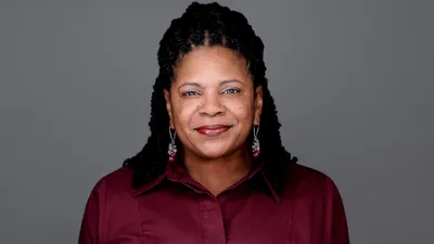 Professional headshot of African-American female with long black twisted braids pulled back, dangling silver and burgundy beaded earrings, warm smile showing teeth, wearing burgundy collared button-down shirt, photographed on grey background in Philadelphia, Pennsylvania by headshot photographer Richard Waine