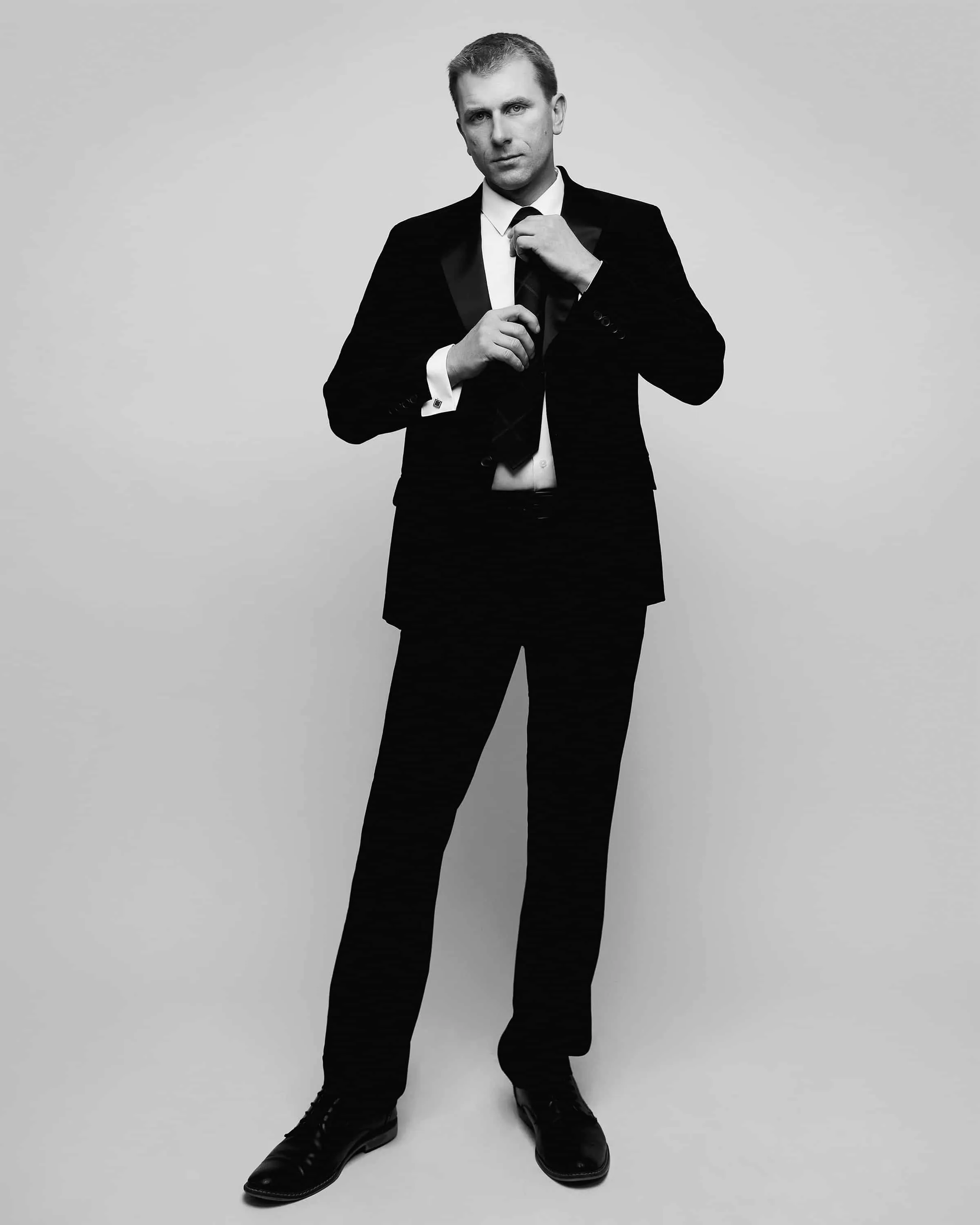 Full-length black and white portrait of male fitness model with short dark hair adjusting black necktie wearing classic black formal suit white dress shirt and black dress shoes in cinematic James Bond style pose photographed against gray studio background by Richard Waine Photography Lancaster PA,  fitness model, professional portraits, professional model portraiture