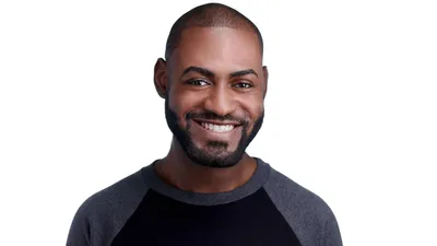 Professional headshot of African-American male with short dark brown hair, trimmed black beard and goatee, wearing grey and black raglan baseball style shirt, bright white smile showing teeth, photographed on seamless white background by Lancaster Pennsylvania headshot photographer Richard Waine