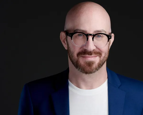 Professional business headshot of psychotherapist with beard and glasses in blue blazer over white shirt, friendly smile and confident facial expression against black studio background, headshot made by Richard Waine in Lancaster Pennsylvania