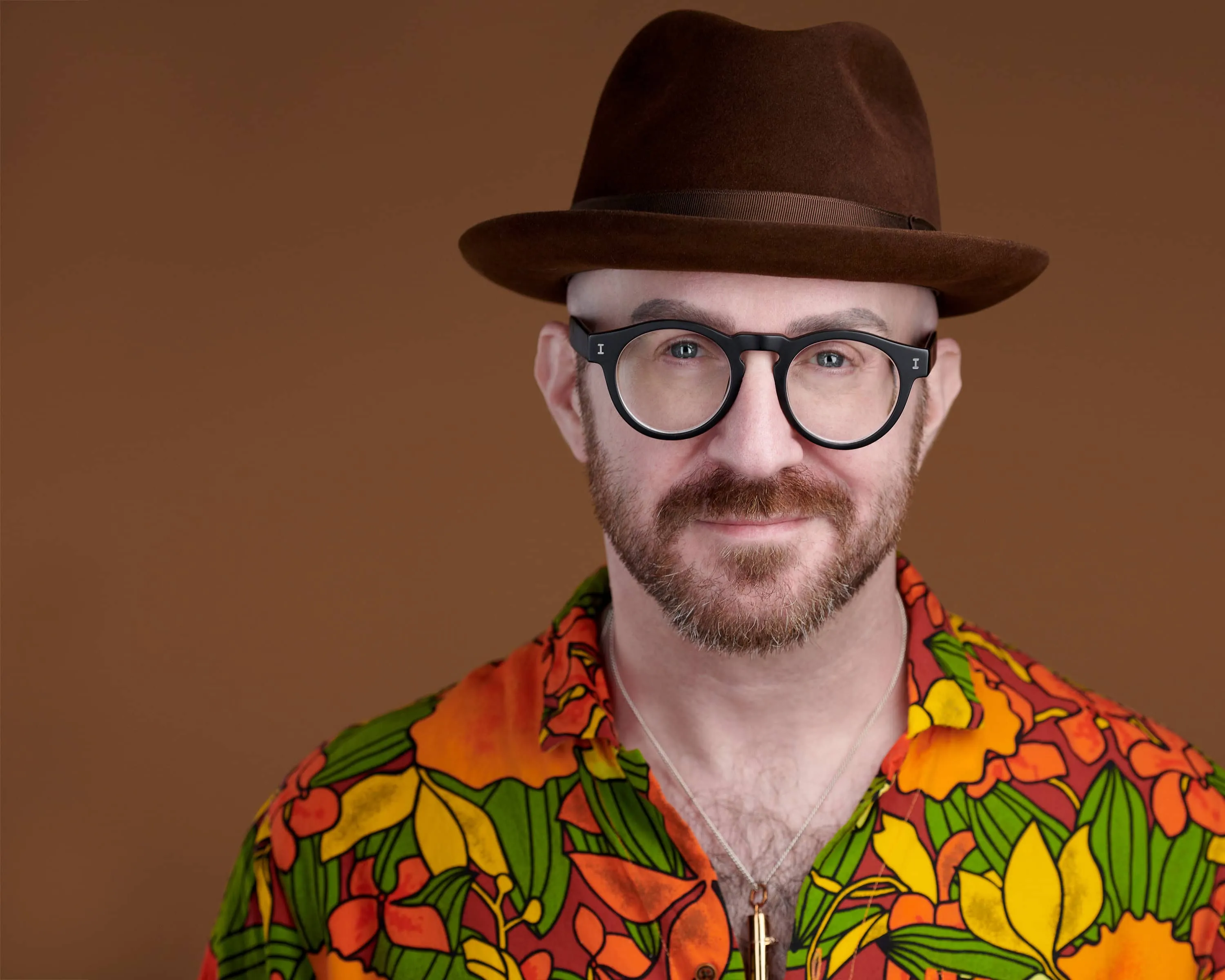 studio headshot of a man wearing a vibrant tropical floral shirt in orange, red, green, and yellow with bold leaf and flower patterns, paired with a chocolate brown felt fedora hat, black round thick-framed glasses, a short groomed beard, and a small pendant necklace, photographed against a warm brown backdrop by Richard Waine Photography in Lancaster, PA