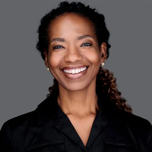 Professional business headshot by Richard Waine of African American woman with curly dark brown hair and radiant smile wearing black v-neck blouse against gray studio background
