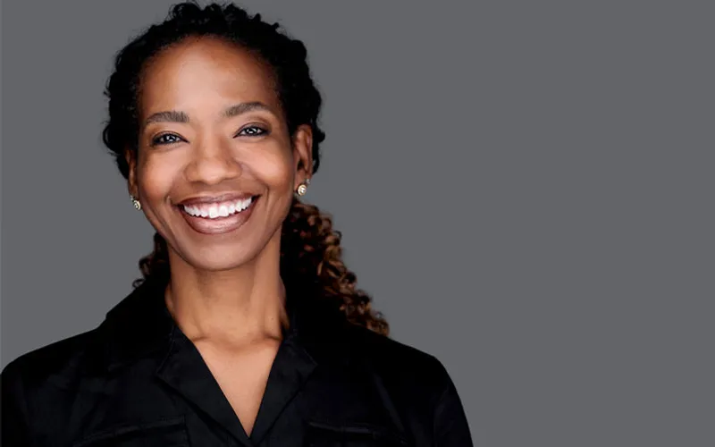 Professional business headshot by Richard Waine of African American woman with curly dark brown hair and radiant smile wearing black v-neck blouse against gray studio background
