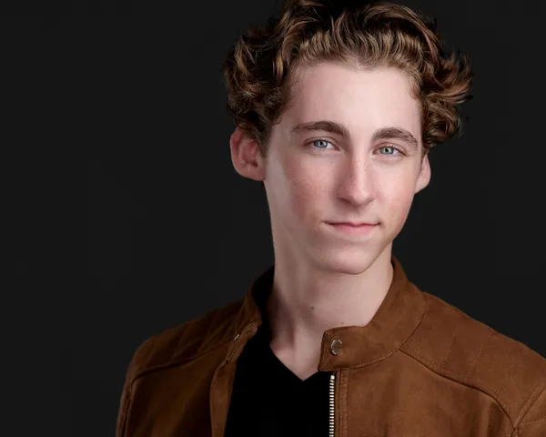 Professional actor headshot of young man with tousled medium-length brown wavy hair and blue eyes with natural light smile wearing tan suede bomber jacket with snap collar over black v-neck shirt photographed against dark charcoal gray studio backdrop with dramatic professional lighting by Richard Waine Photography actor headshot photographer Lancaster Philadelphia Pennsylvania Newark Wilmington Delaware, acting headshots lancaster, headshots for actors, Lancaster studio headshots