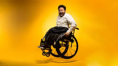Philadelphia photographer Jaleel King creative portrait in black wheelchair with short natural hair and full beard, wearing tan beige button-down shirt with black pants, mouth open in excited joyful expression, photographed against vibrant yellow orange gradient background, disability representation in professional photography industry
