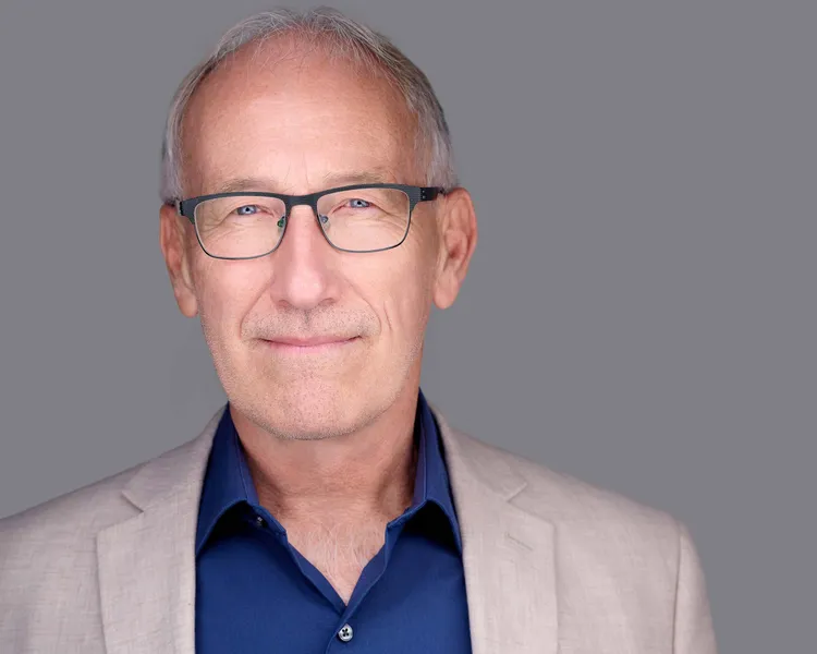 Professional business headshot of man with short silver-gray hair and trimmed gray stubble wearing dark gray rectangular frame eyeglasses tan linen blazer over navy blue open-collar dress shirt photographed against neutral gray studio background by Richard Waine Photography