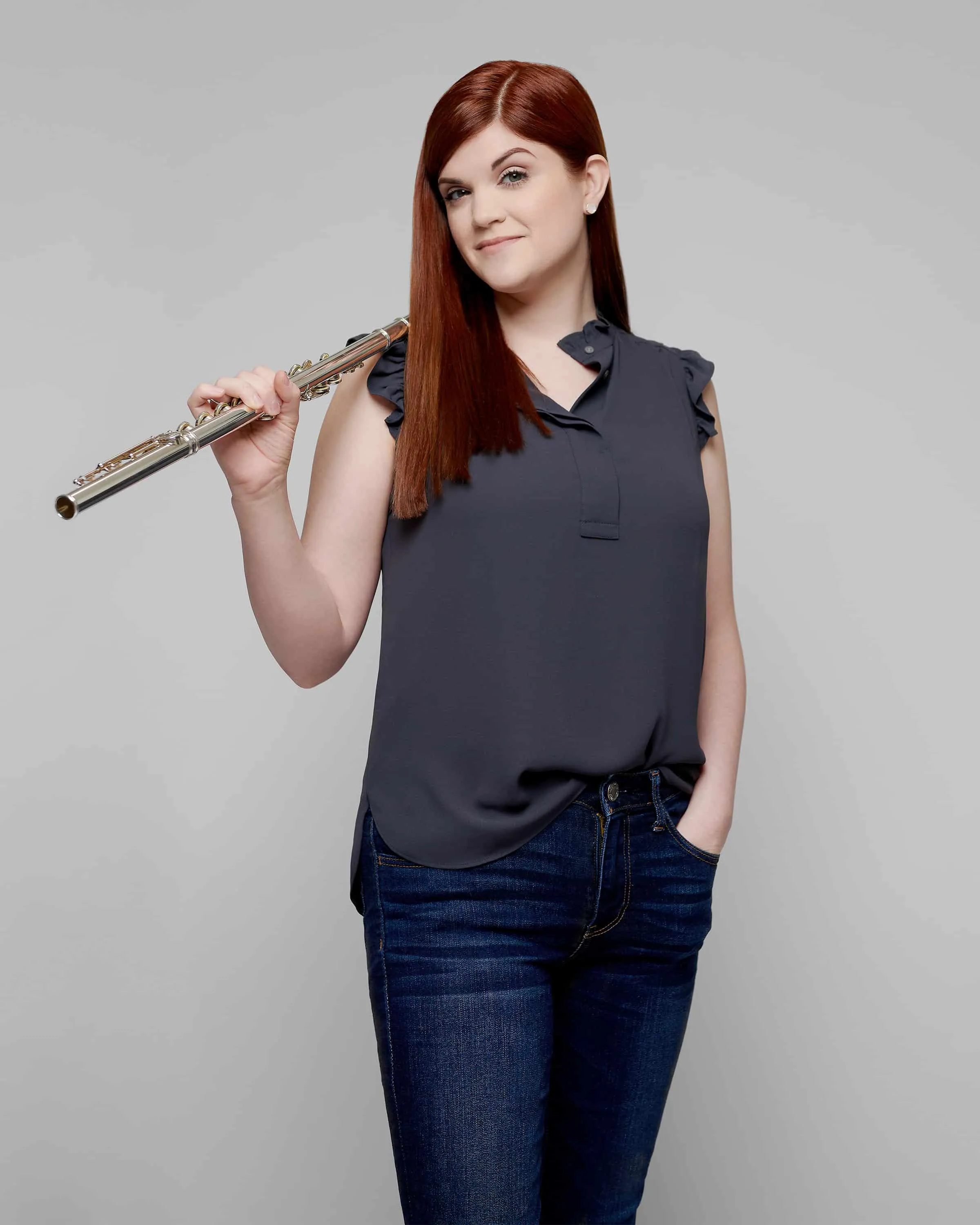 Three-quarter length portrait of female flutist with long straight auburn red hair holding silver flute wearing charcoal gray sleeveless ruffle shoulder blouse with dark wash blue jeans photographed against neutral gray studio background by Richard Waine Photography Lancaster Pennsylvania