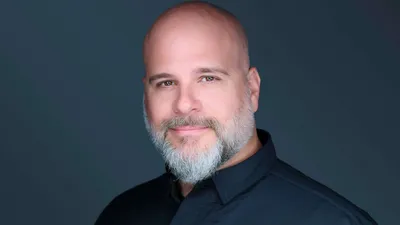 Sheboygan Wisconsin photographer Jon Erlien professional headshot with bald head, full grey beard, wearing black collared shirt photographed on dark teal grey background, interview subject specializing in headshots branding portraits and commercial photography