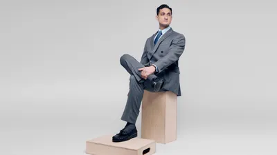 Professional man in a grey double-breasted suit, light blue shirt, and navy tie sitting on a wooden apple box against a neutral grey background