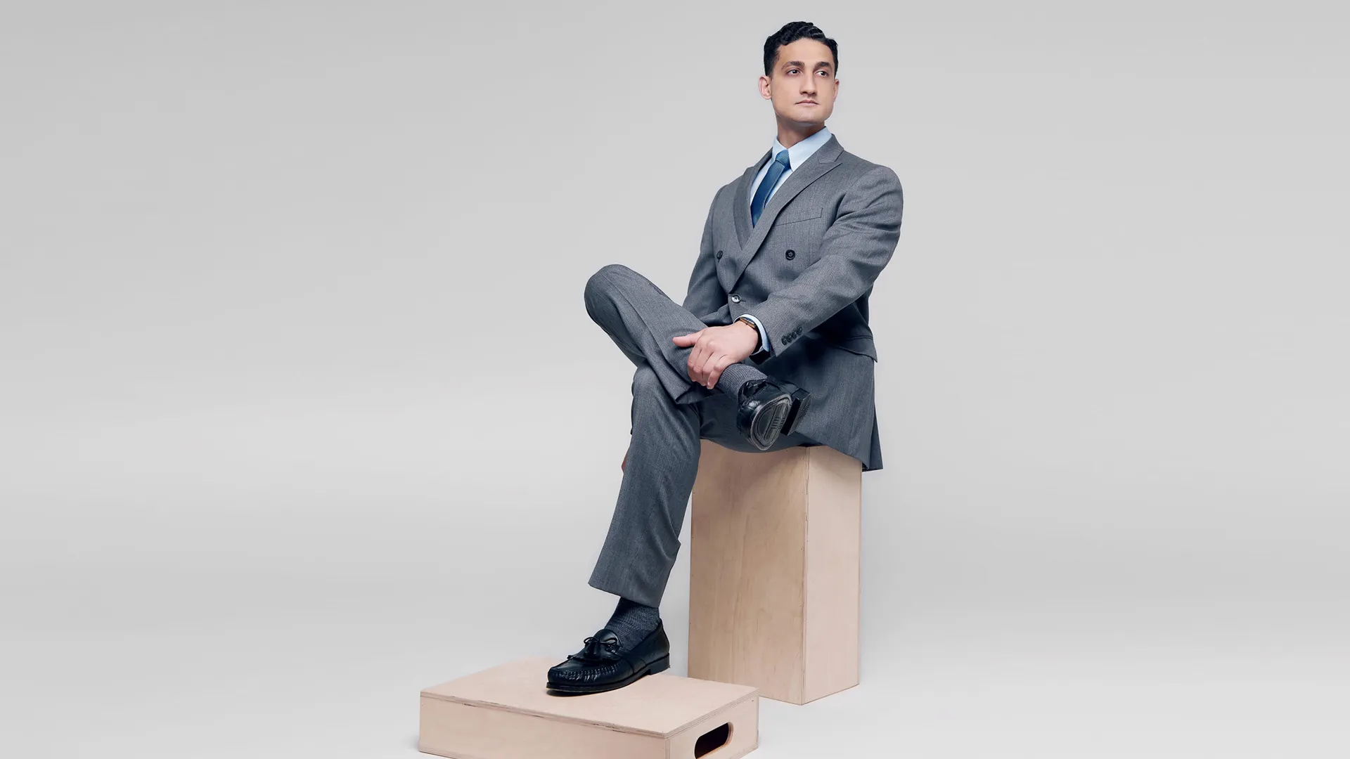 Professional man in a grey double-breasted suit, light blue shirt, and navy tie sitting on a wooden apple box against a neutral grey background