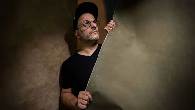 New York photographer Pete Coco creative self-portrait with oversized round tortoise-shell eyeglasses, grey beard stubble, all black outfit peeking around textured tan beige wall with dramatic shadow lighting, interview subject specializing in headshots portraits and music photography