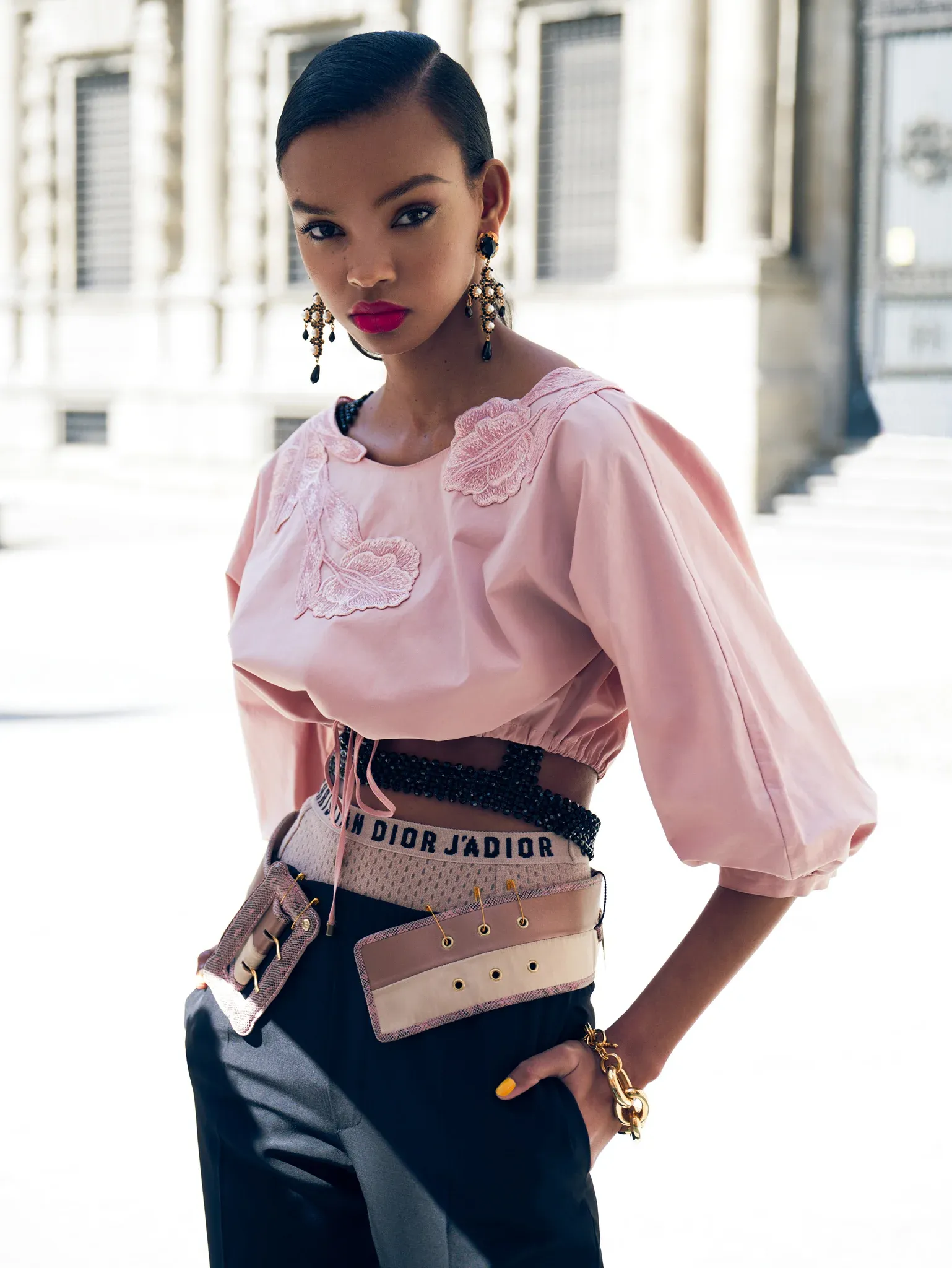 High fashion street style portrait of Black female model with sleek slicked hair, bold magenta lips, dramatic chandelier earrings, wearing pink rose-embroidered cropped blouse with Dior designer belt and black pants, confident direct gaze, bright urban European architecture background, professional editorial retouching by Pratik Naik for photographer Dennison Bertram
