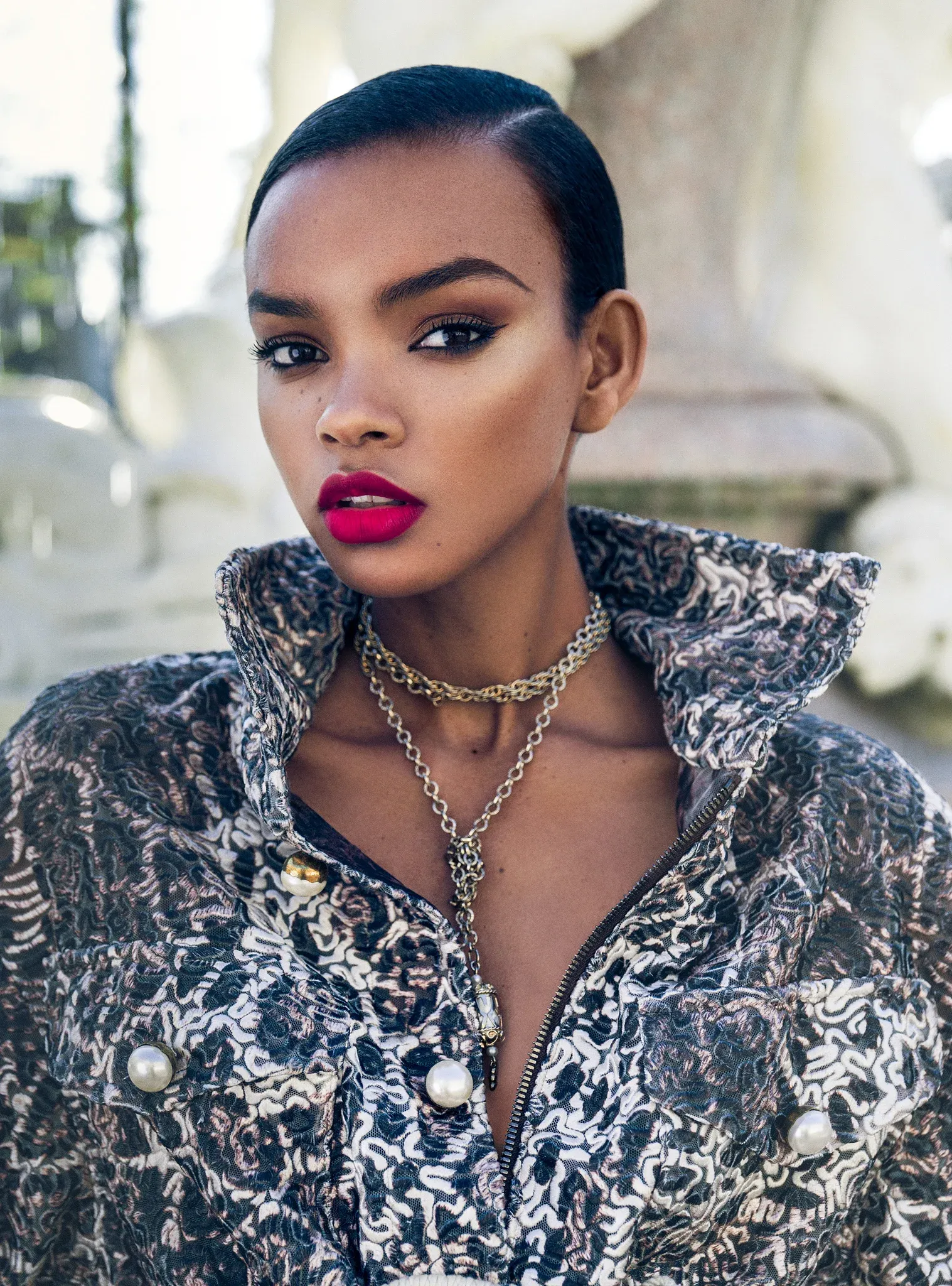 High fashion editorial portrait of Black female model with sleek slicked short hair, dramatic bold fuchsia pink lips, striking eye makeup, wearing ornate black and white patterned structured jacket with pearl buttons and statement chain necklaces, bright outdoor architecture background, professional beauty retouching by Pratik Naik for photographer Dennison Bertram