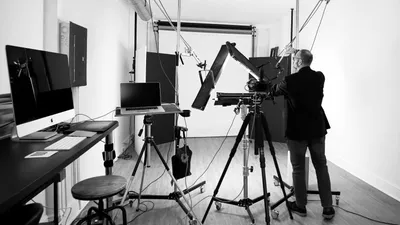 Black and white behind-the-scenes photograph of photographer Richard Waine in dark business suit adjusting professional studio lighting equipment including large rectangular light panels and modifiers mounted on tripod stands in bright white photography studio with tethered workstation showing dual monitors laptop and round stool visible demonstrating professional headshot session setup at Richard Waine Photography Lancaster Pennsylvania