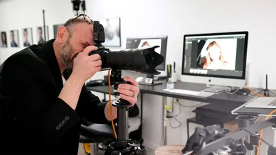 Behind the scenes photography session showing photographer Richard Waine in black blazer with brown hair and beard looking through viewfinder of Nikon DSLR camera mounted on orange and black tripod, computer monitors displaying headshot images on desk in background, Lancaster, Pennsylvania headshot photography studio workspace