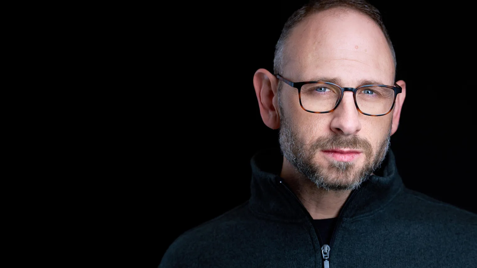 Professional headshot of Richard Waine Lancaster Pennsylvania headshot photographer with short brown hair receding hairline and trimmed brown and gray beard wearing tortoise-shell frame eyeglasses and dark gray quarter-zip fleece pullover photographed against black studio background with dramatic studio lighting by colleague photographer Sanjeev Singhal