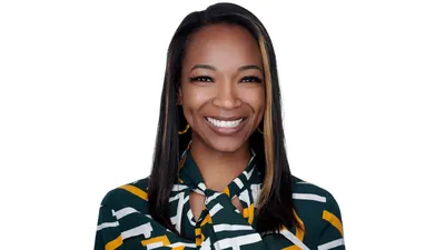 Professional corporate headshot of African American woman with medium brown skin tone and long straight dark hair with blonde and caramel highlights framing face wearing dark forest green blouse with bold white and gold geometric striped pattern with gold mustard colored hoop earrings showing genuine warm expression photographed against seamless white background with professional high-key lighting by Richard Waine Photography Lancaster Pennsylvania