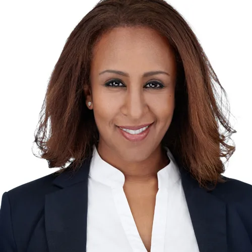 Professional business headshot of African-American woman with shoulder-length reddish-brown wavy hair and warm smile wearing navy blue blazer over white collared blouse with diamond stud earrings photographed against seamless white backdrop by Richard Waine