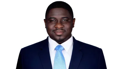Professional corporate headshot of African American man with deep brown skin tone short cropped dark hair and neatly trimmed goatee and mustache facial hair with warm brown eyes and confident professional expression wearing navy blue business suit jacket with notched lapels over crisp white dress shirt with spread collar and light blue silk tie with small dotted pattern photographed against seamless white studio background with professional high-key lighting by Richard Waine Photography Lancaster Pennsylvania