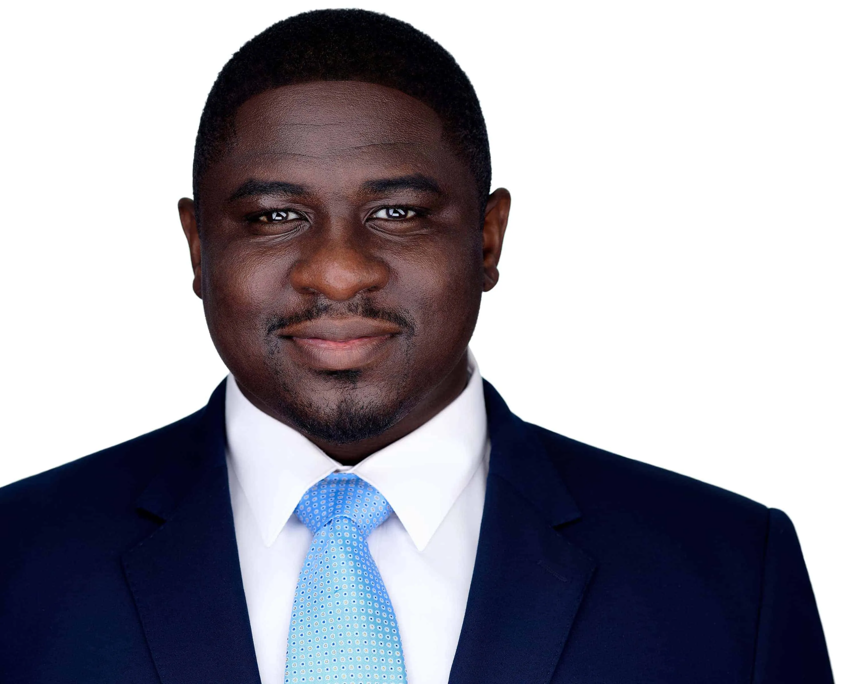 Professional corporate headshot of African American man with deep brown skin tone short cropped dark hair and neatly trimmed goatee and mustache facial hair with warm brown eyes and confident professional expression wearing navy blue business suit jacket with notched lapels over crisp white dress shirt with spread collar and light blue silk tie with small dotted pattern photographed against seamless white studio background with professional high-key lighting by Richard Waine Photography Lancaster Pennsylvania