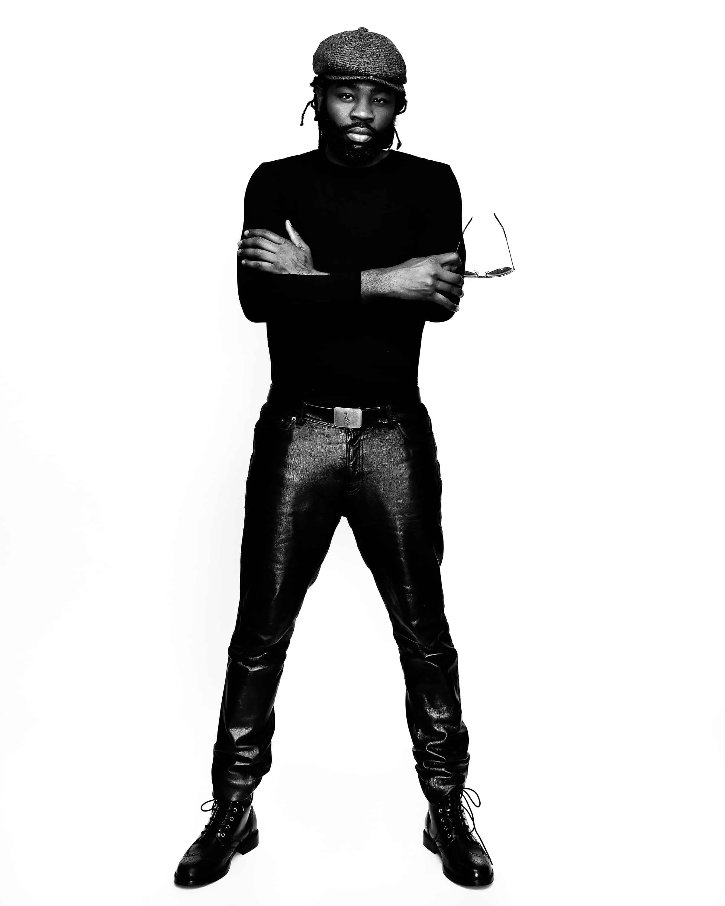 Full-length black and white portrait of male musician with locs tucked under charcoal newsboy cap holding sunglasses dressed in all black outfit with crew neck sweater black leather pants silver belt buckle and lace-up boots photographed against white studio background by Richard Waine